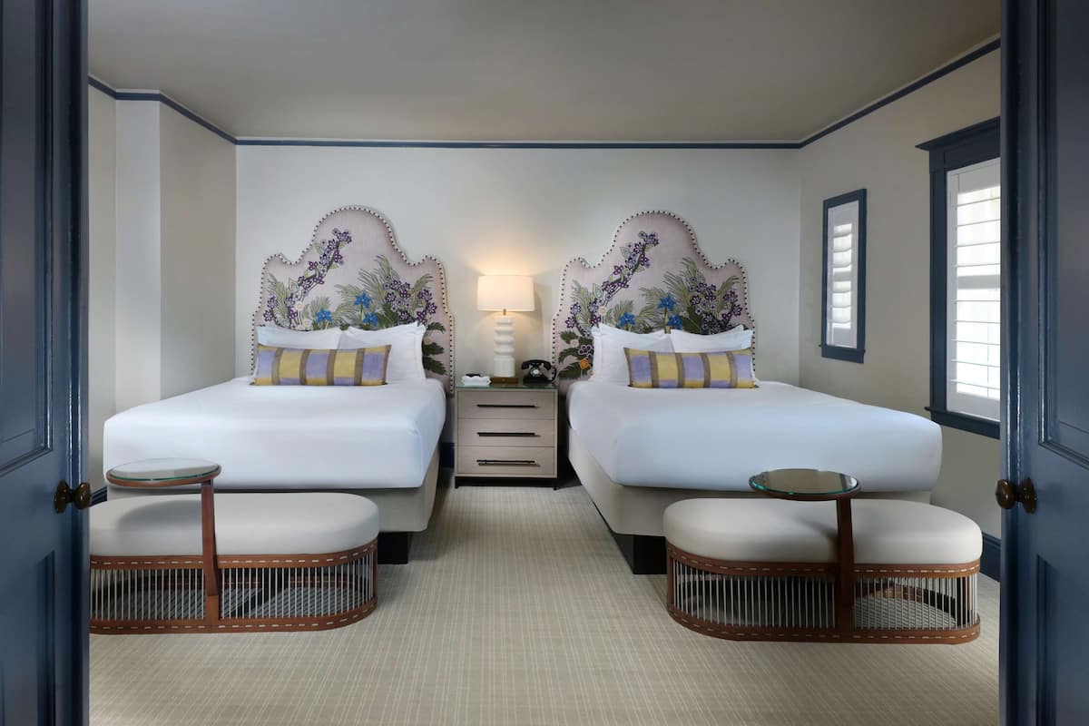 Une chambre d’hôtel avec deux lits, une petite table, une lampe et une fenêtre avec des stores.