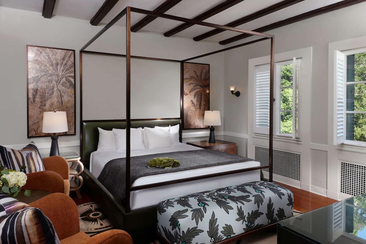 Un lit à baldaquin avec du linge de lit blanc, un plaid vert et un pouf orné d’un motif floral. Il y a deux tables de chevet avec des lampes, une grande fenêtre avec des volets et une vue sur la verdure à l’extérieur.