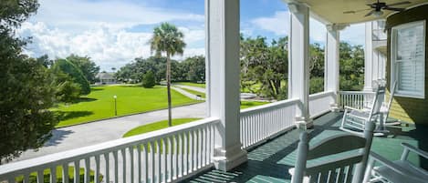 A covered porch with white railings and green flooring, overlooking a well-maintained lawn and a road.