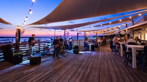 Una terraza de madera con luces colgantes y un escenario para música en vivo, con vistas a una masa de agua.