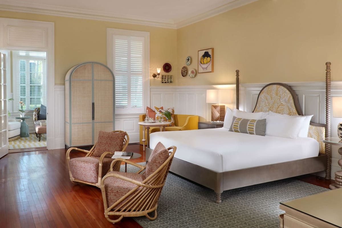 Une chambre à coucher avec un grand lit, deux fauteuils en osier, une petite table et une fenêtre avec des stores.