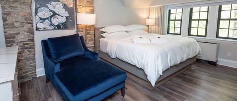 Un dormitorio con una pared de acento de piedra, una cama grande con ropa de cama blanca, una silla azul y un cuadro de flores.