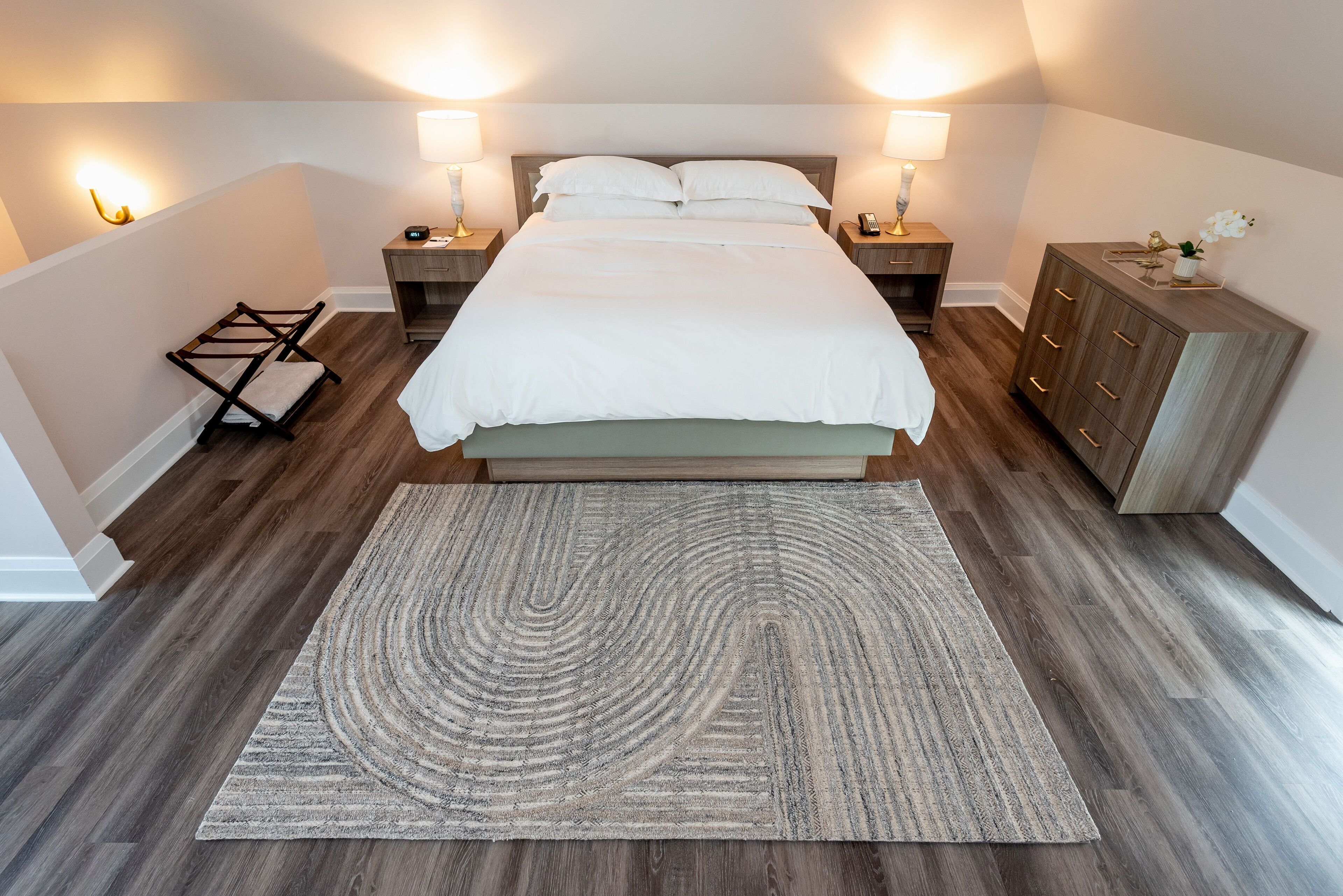 Un dormitorio ordenado con una cama grande, lámparas de noche, una mesita de madera, un aparador y un tapete estampado.
