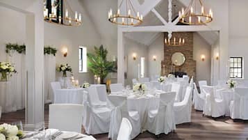 Un salón de banquetes con mesas redondas, sillas blancas, candelabros y una pared de piedra.