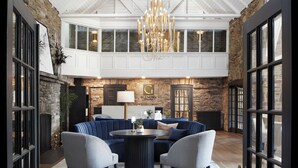A spacious living area with a large blue sofa, two chairs, a round coffee table, and a chandelier. There is a stone wall with a fireplace and a large window above the sofa.