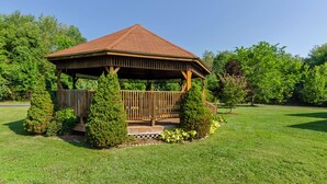 Un belvédère en bois, surmonté d’un toit en tuiles, entouré de pelouse bien entretenue et d’arbres.