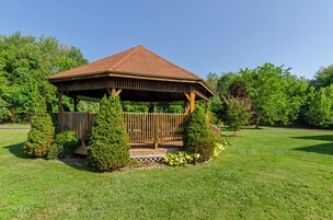 Um gazebo de madeira com telhado de telhas, cercado por gramado bem cuidado e árvores.