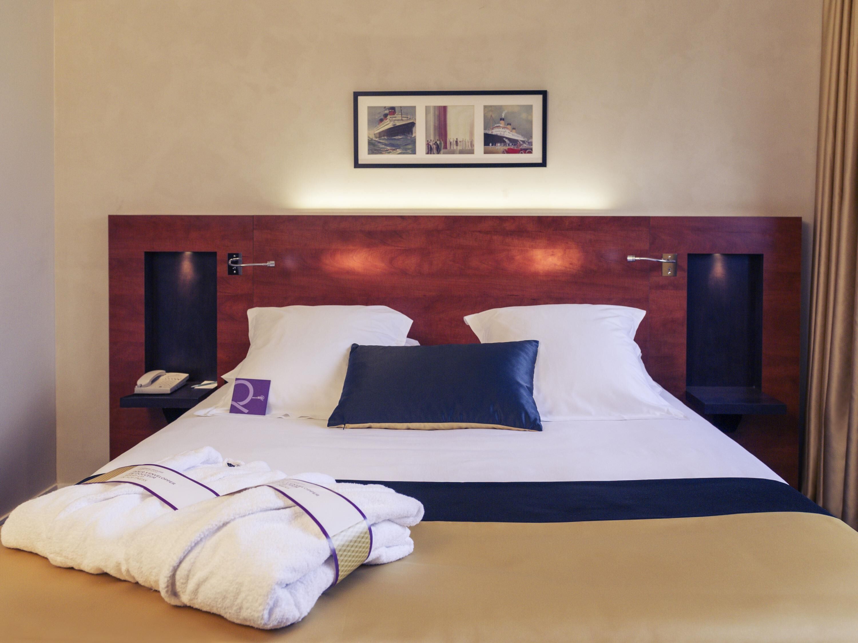 Habitación de hotel con una cama grande, dos mesitas de noche, un paragüero y un cuadro enmarcado sobre la cama, además de un juego de toallas sobre la cama.
