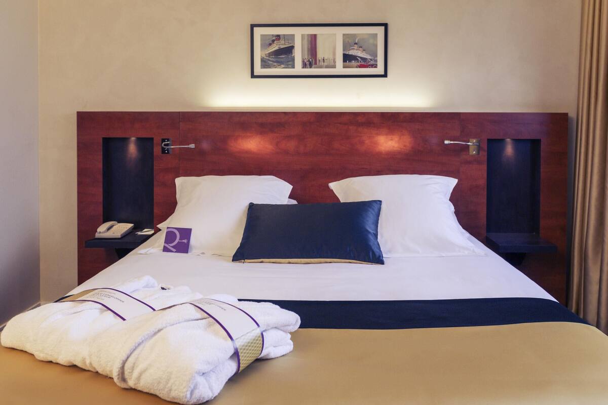 Habitación de hotel con una cama grande, dos mesitas de noche, un paragüero y un cuadro enmarcado sobre la cama, además de un juego de toallas sobre la cama.