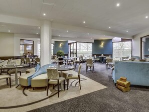 Un amplio vestíbulo de hotel con varias zonas de estar, incluyendo mesas redondas y sofás, y ventanas grandes que proporcionan luz natural.