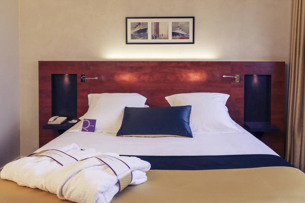 A hotel room with a bed, pillows, a bedside table with a phone, a framed picture above the bed, and a towel on the floor.