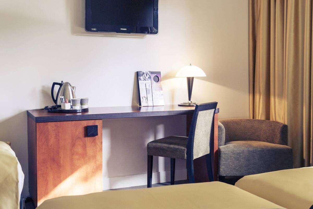 Una habitación de hotel con escritorio, una silla, televisor montado en la pared y lámpara.