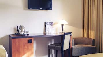 A hotel room with a desk, a chair, a television mounted on the wall, and a lamp.