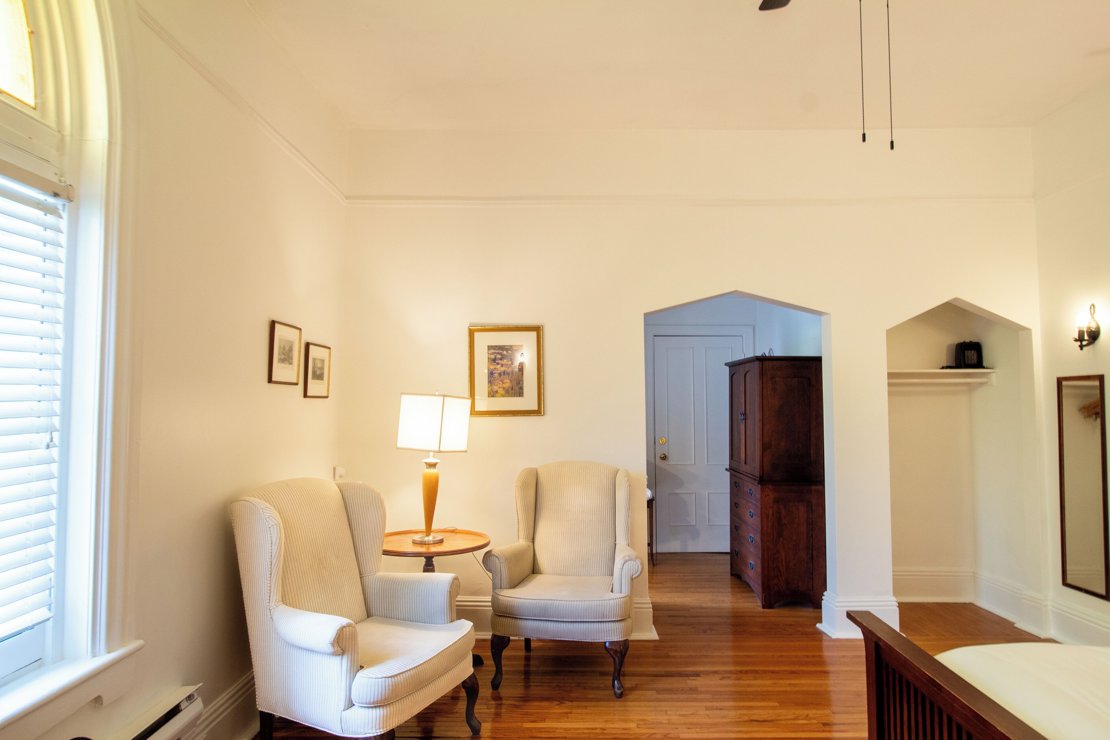 A room with a wooden floor, two armchairs, a table with a lamp, a door, and a mirror on the wall.