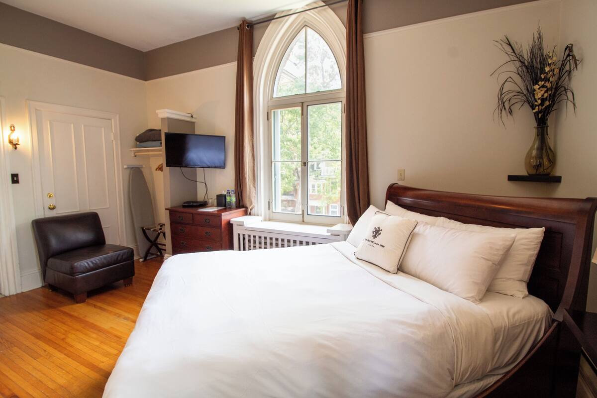 A bedroom with a large bed, a television on a stand, a chair, a radiator, and a window with curtains.