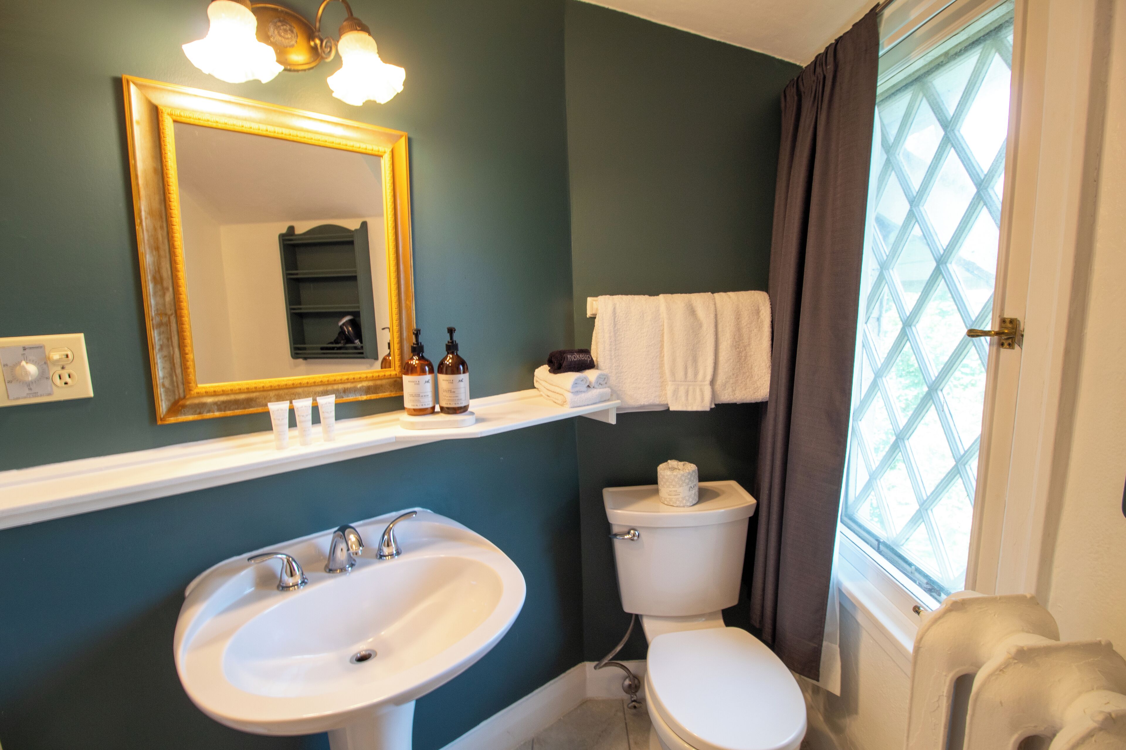 Une salle de bain avec un lavabo blanc, des toilettes, un miroir et une fenêtre avec des rideaux.