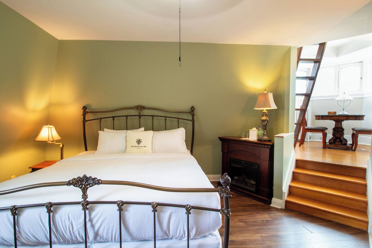 A bedroom with a four-poster bed, a nightstand with a lamp, a fireplace, and a staircase leading to an upper level.
