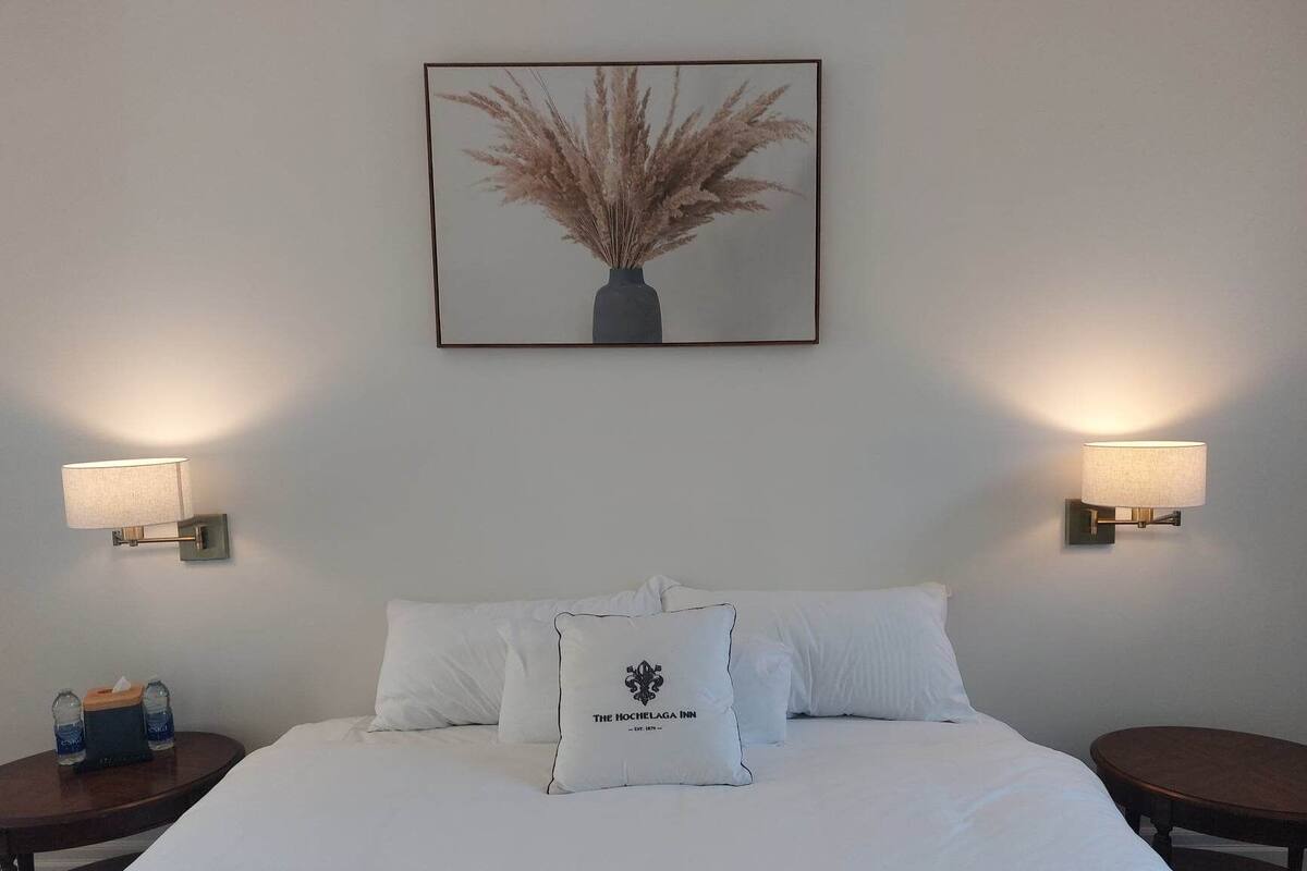 A hotel room with a large bed, two bedside tables, a wall-mounted lamp, and a framed picture above the bed.