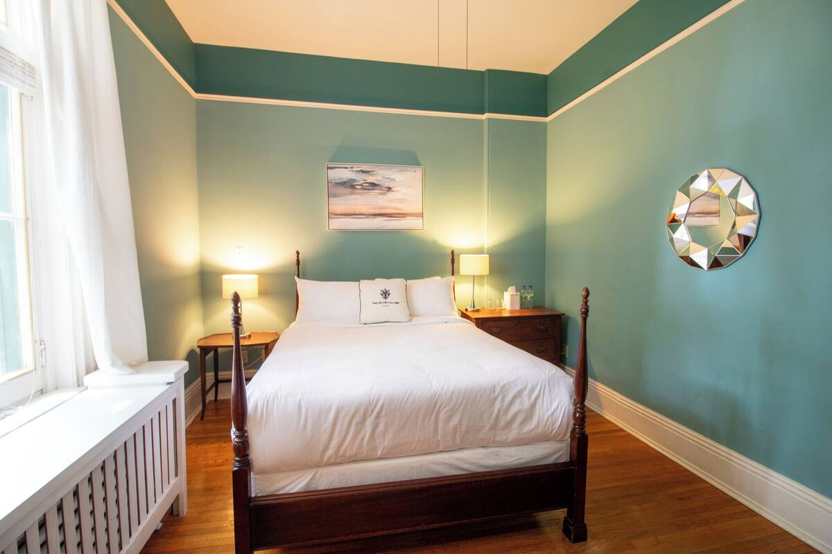 A bedroom with a wooden four-poster bed, two bedside tables with lamps, a wall-mounted mirror, and a painting above the bed.
