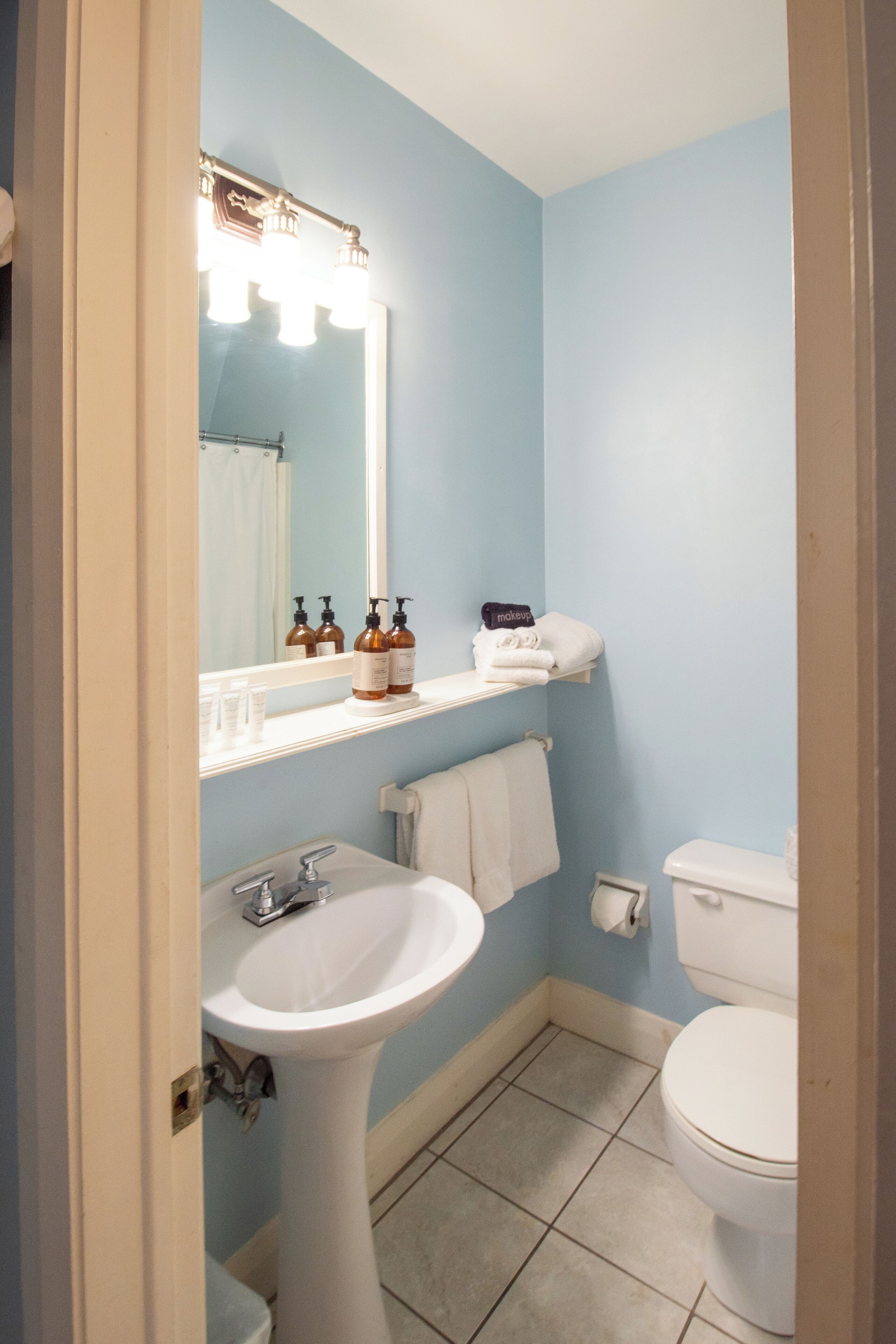 A bathroom with a pedestal sink, a white toilet, and a mirror with lights. There are toiletries on the shelf above the sink.