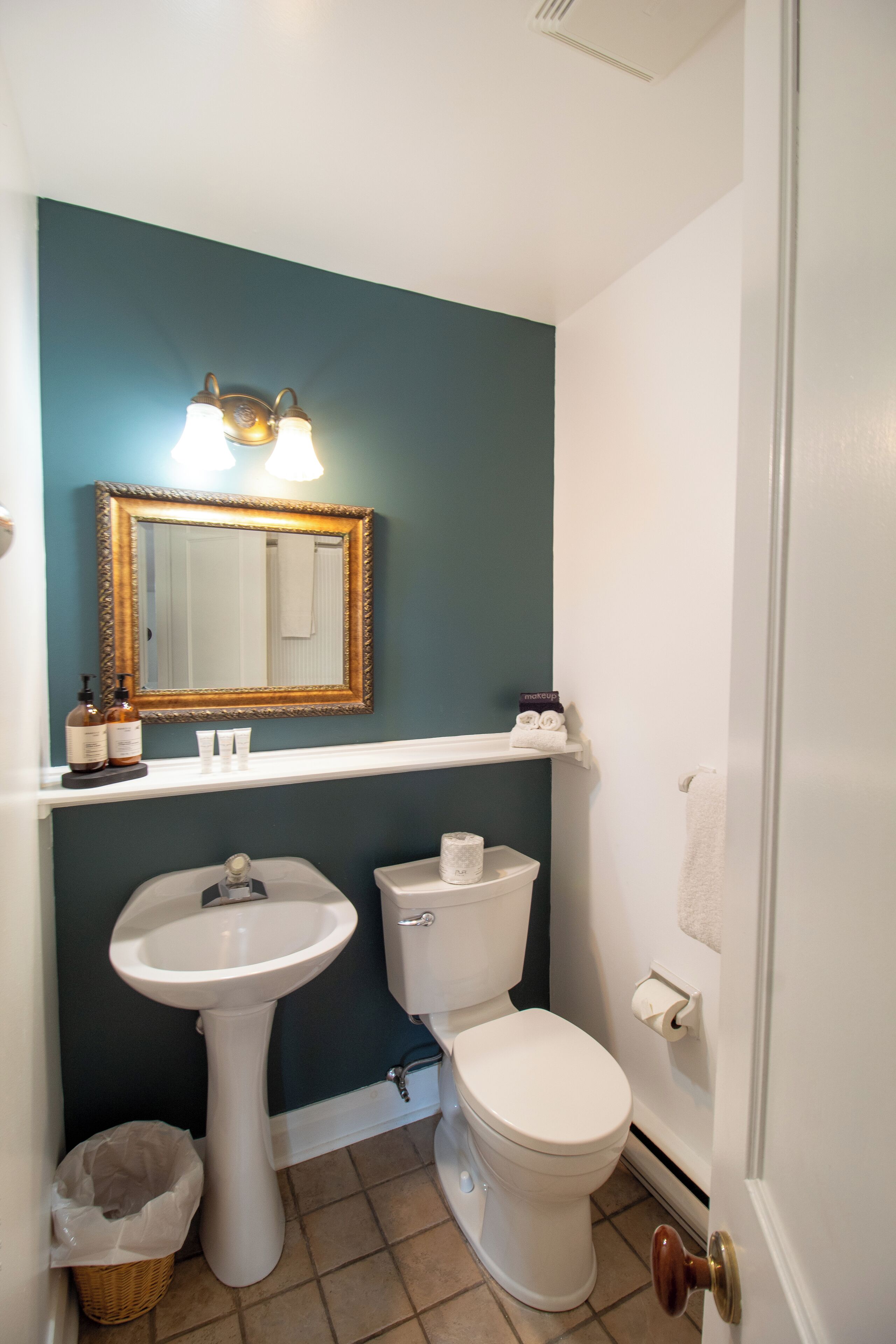 Ein Badezimmer mit einem weißen Waschbecken, einer Toilette und einem Spiegel mit Beleuchtung. Ein Korb und ein Handtuchhalter sind ebenfalls vorhanden.