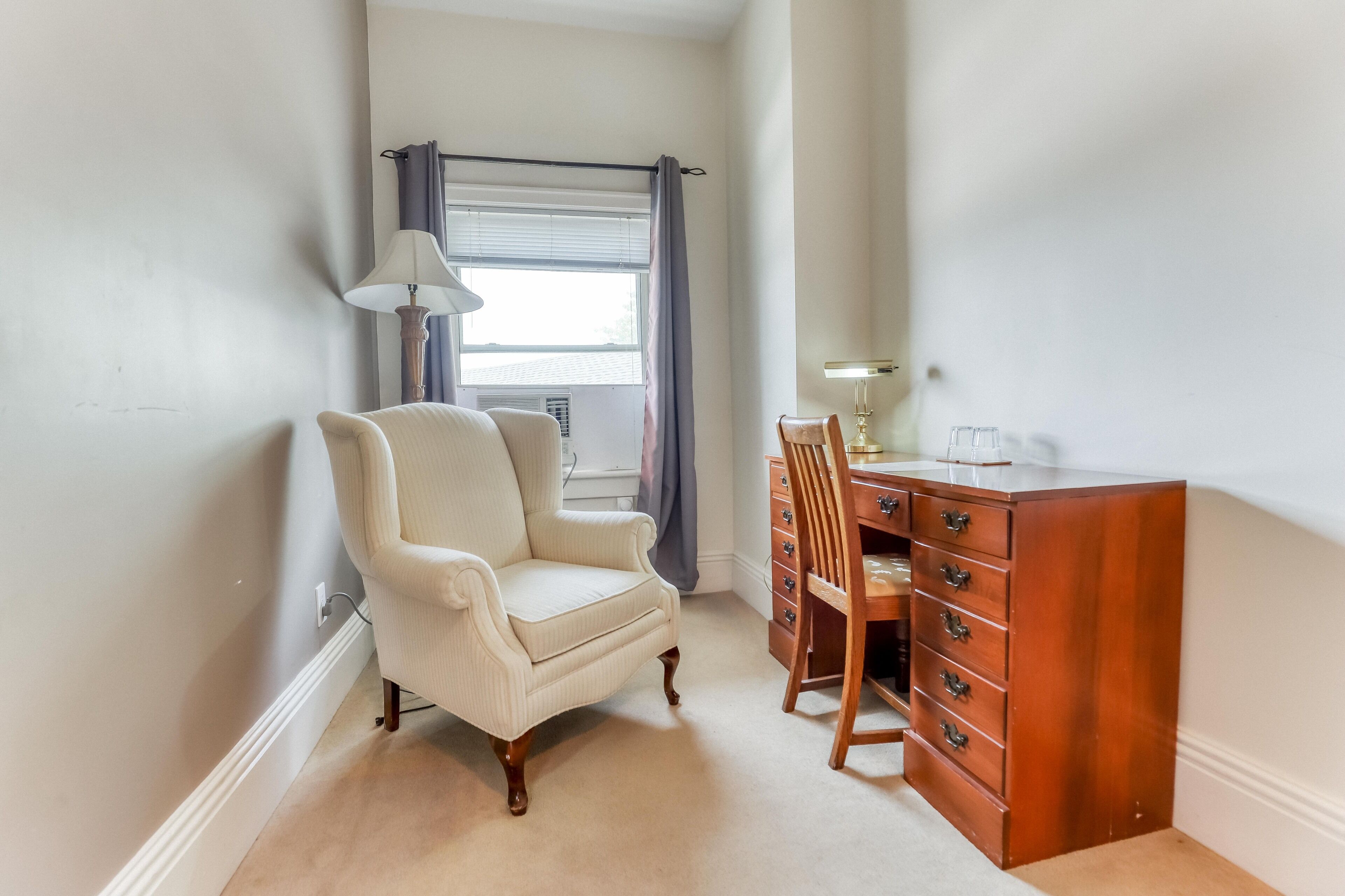 A corner room with a white armchair, a wooden desk with drawers, a lamp, and a window with curtains.