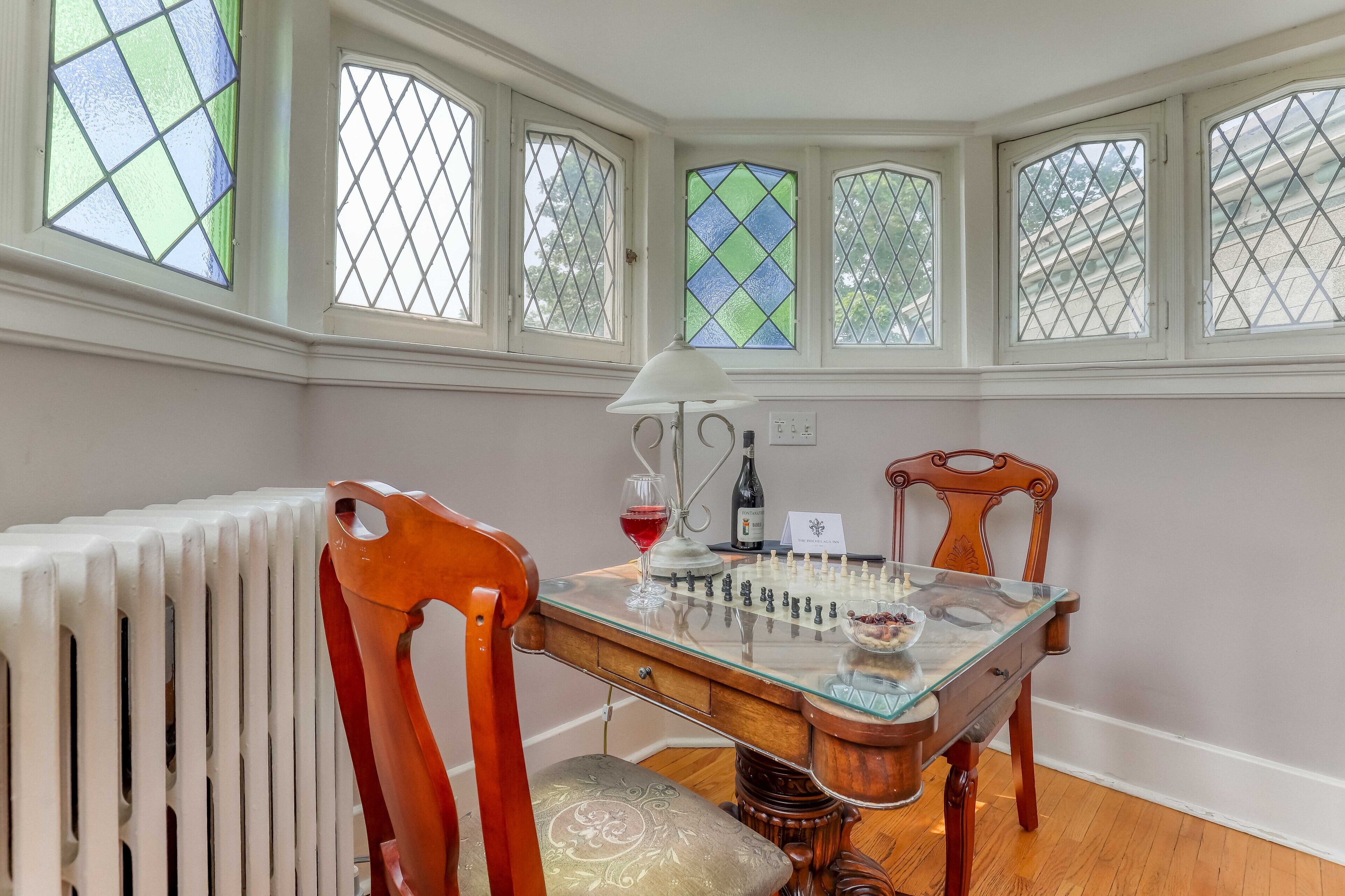 A room with a glass-top table, a chessboard, a wine glass, and a bottle. There are four windows with stained glass panes.
