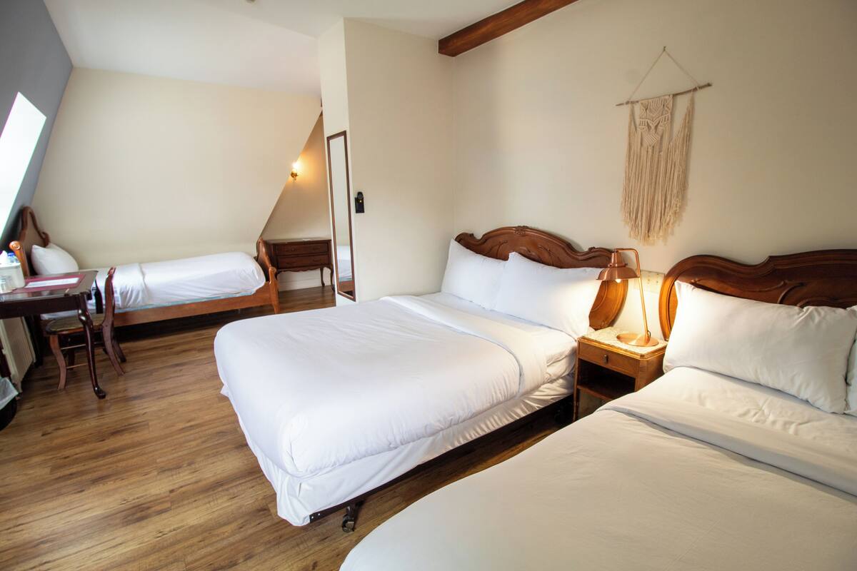 A hotel room with two beds, a wooden floor, a desk with a chair, a lamp, and a wall-mounted macrame decoration.