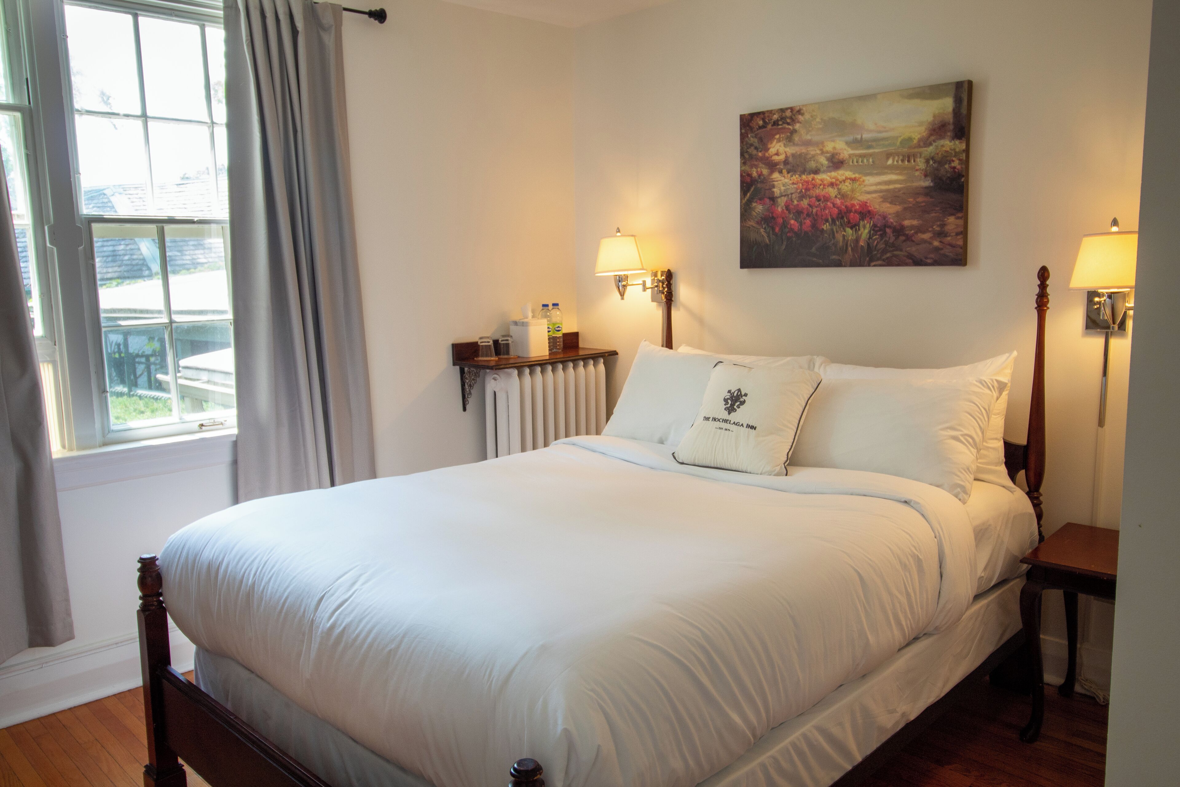 Un lit bien fait, avec du linge de lit blanc, une table de chevet avec une lampe, un radiateur et une fenêtre avec des rideaux.