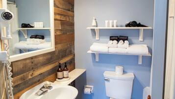 Un baño con pared de madera, un lavamanos blanco, un inodoro y estantes con artículos de aseo.