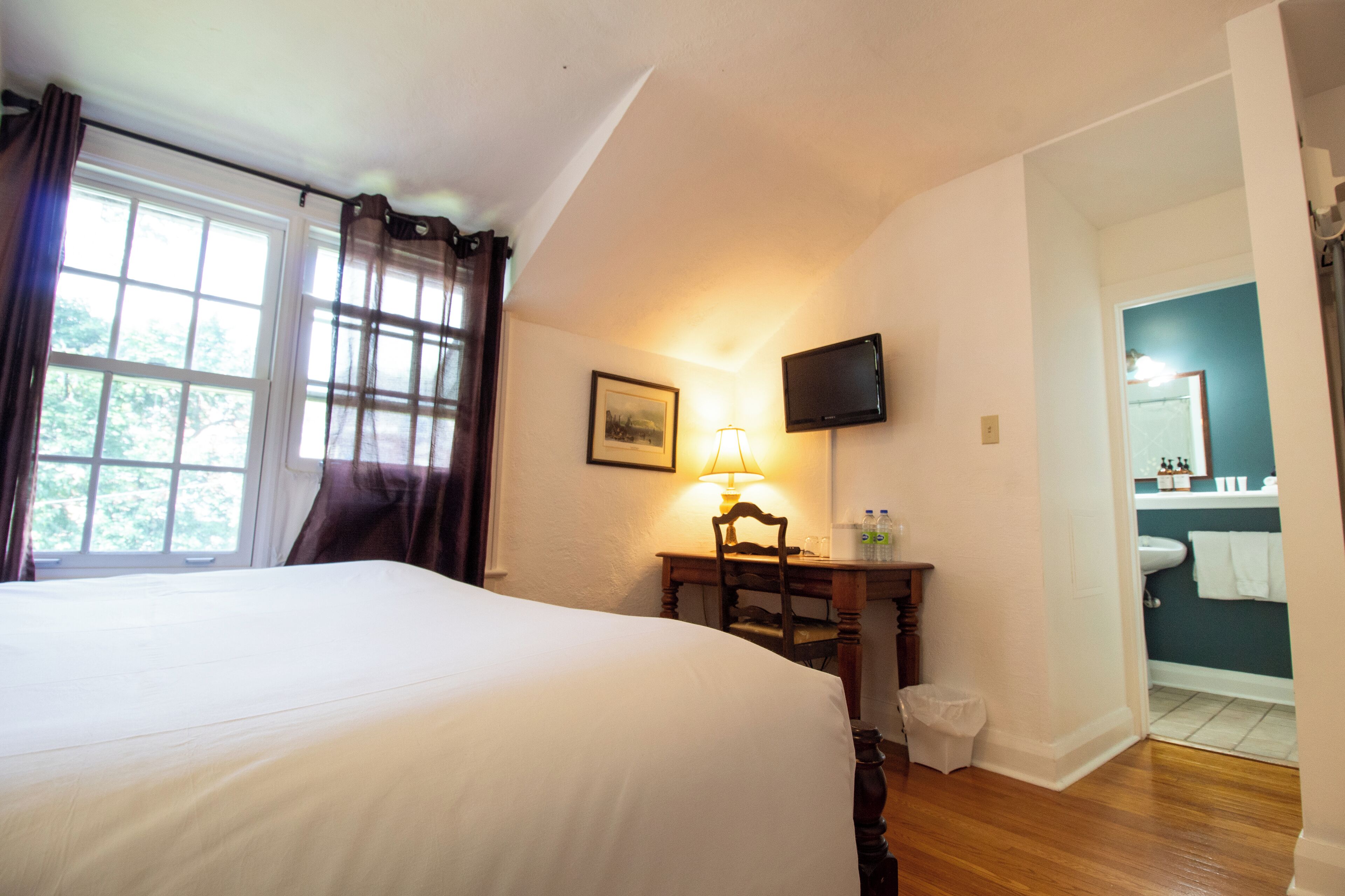 A hotel room with a large bed, a desk with a lamp, a television, and a view of the bathroom.