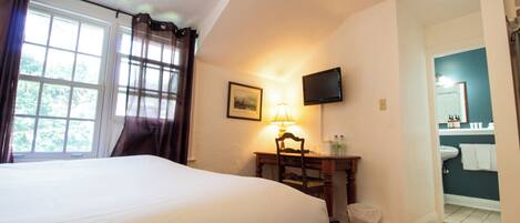 A hotel room with a large bed, a desk with a lamp, a television, and a view of the bathroom.