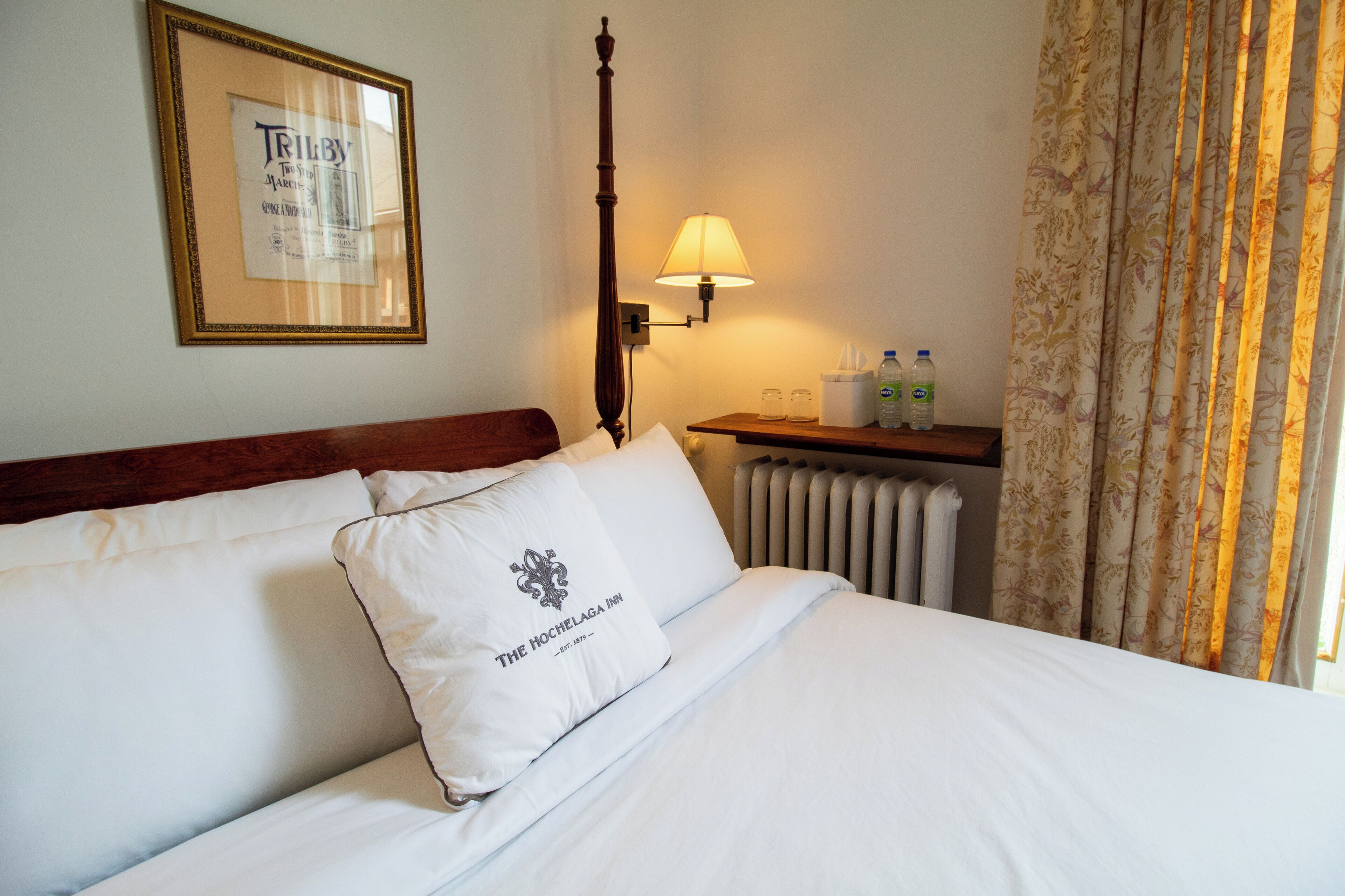 Una habitación de hotel con una cama, un menú enmarcado en la pared, una mesita de noche con artículos de aseo y un radiador.