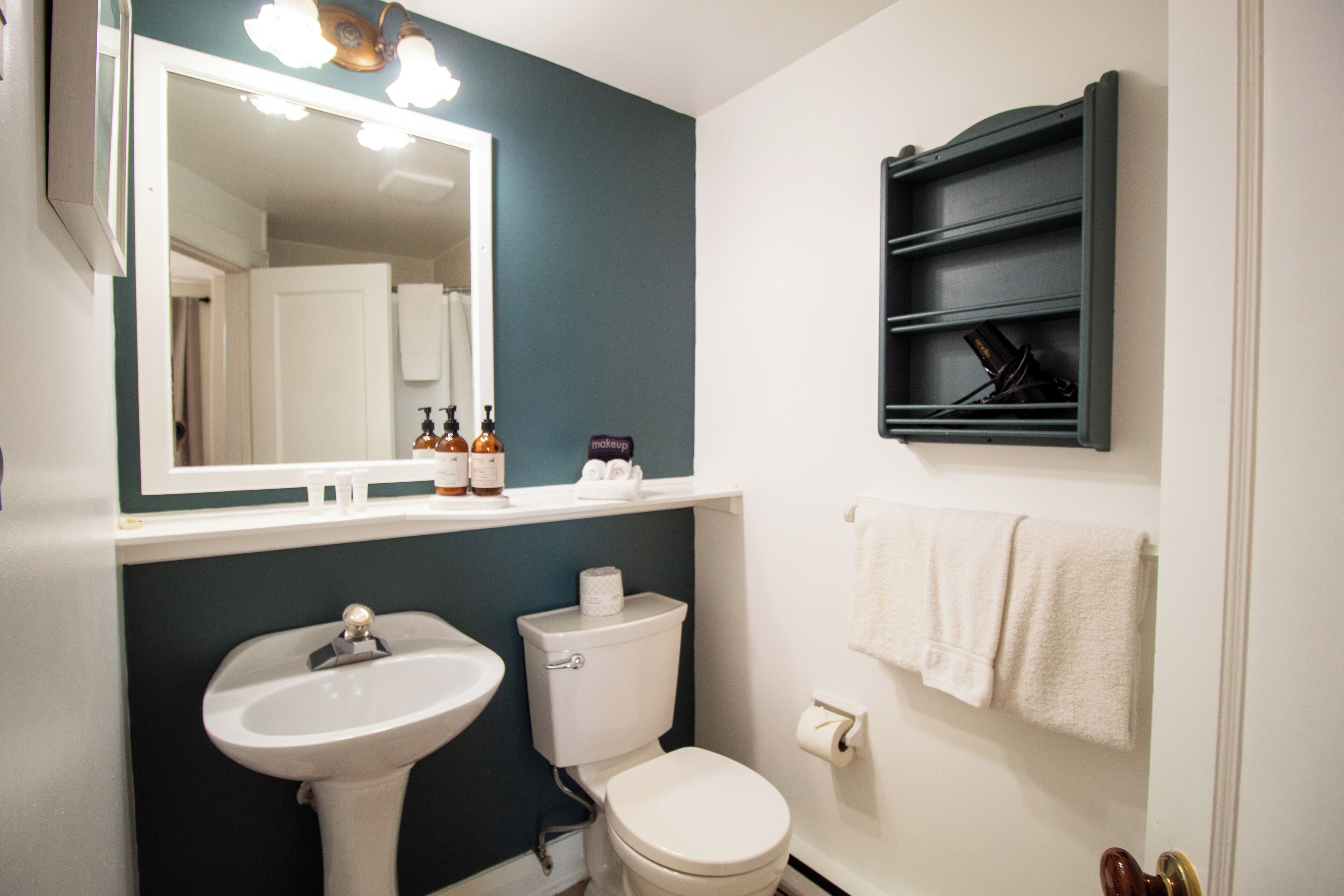 A bathroom with a white sink, a toilet, a mirror, and a shelf with toiletries.