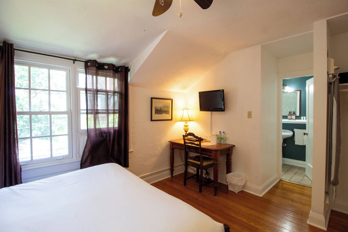 A bedroom with a bed, a desk with a chair, a lamp, a television, and a bathroom visible through an open door.