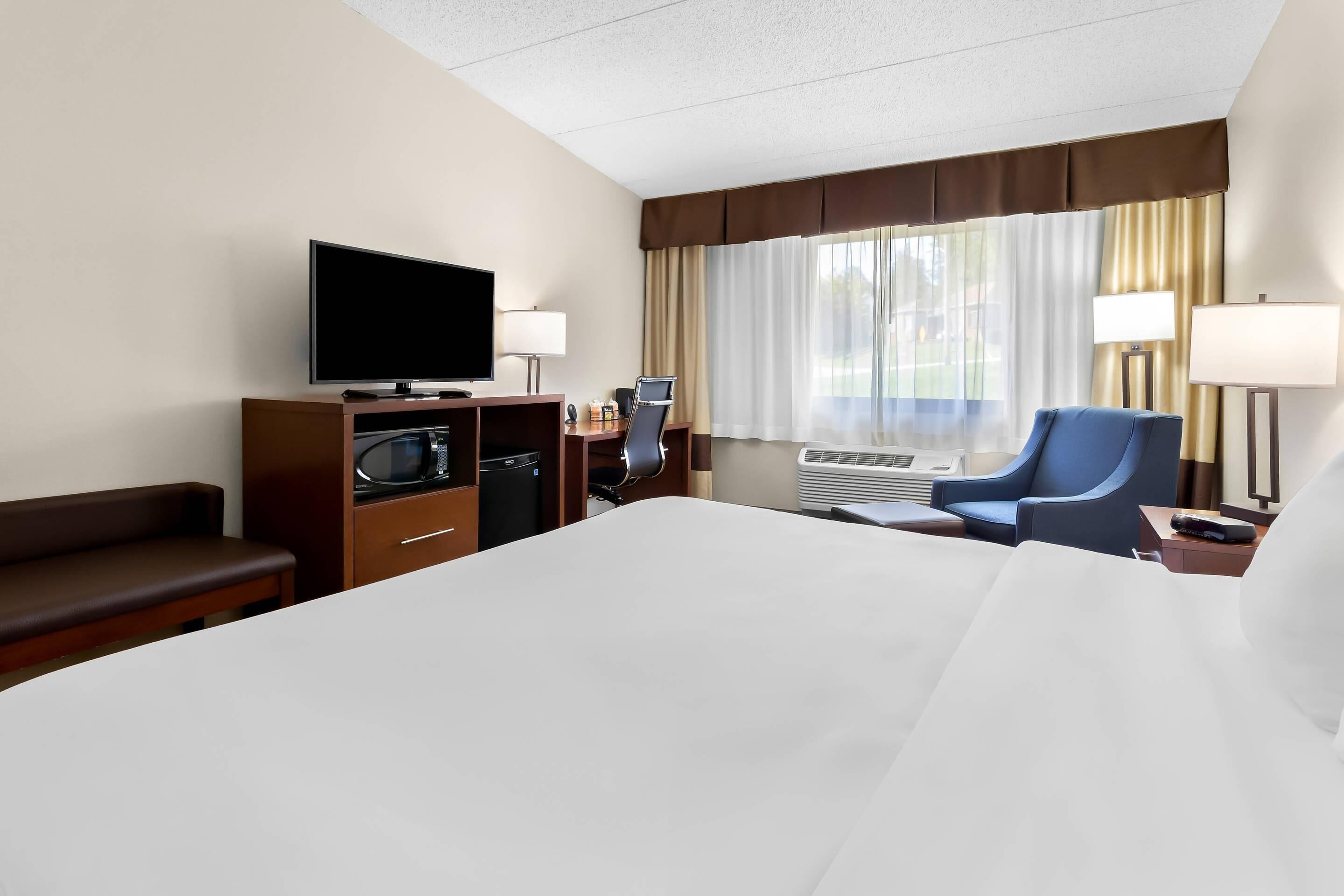 A hotel room with a large bed, a flat-screen TV, a desk with a computer, a microwave, a chair, and a window with curtains.