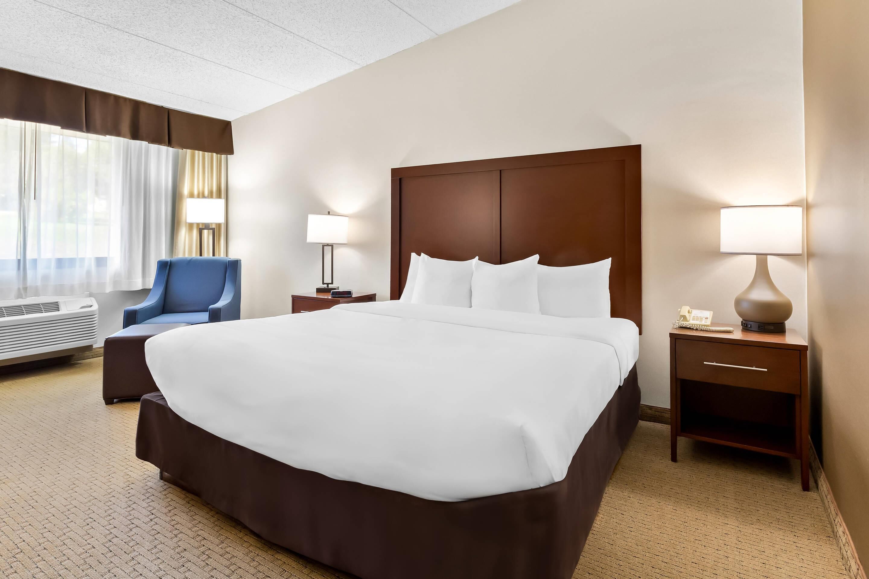 A hotel room with a large bed, a chair, a bedside table with a lamp, and an air conditioning unit.