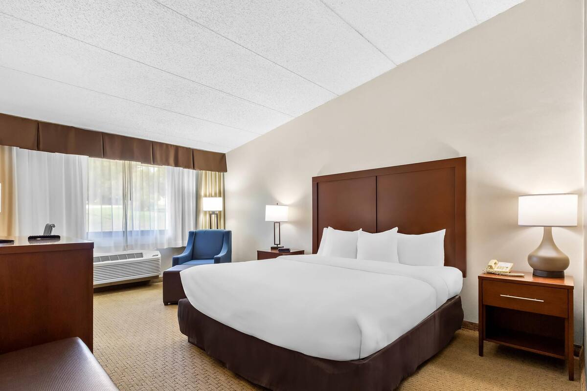 A hotel room with a large bed, a desk, a chair, a bedside table with a lamp, and a window with curtains.