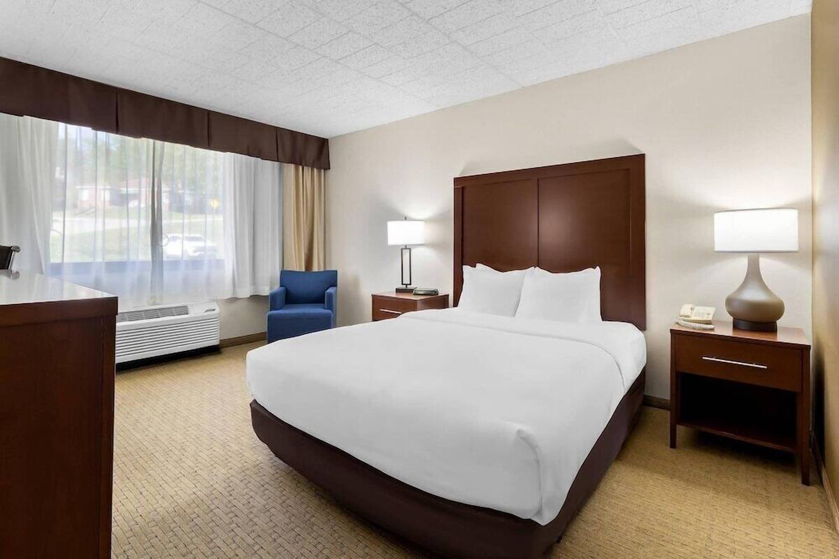 A hotel room with a large bed, a desk, a chair, a bedside table with a lamp, and a window with curtains.