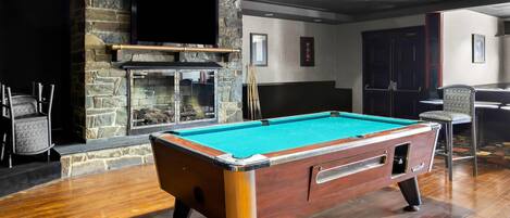 A pool table in a room with a stone fireplace, wooden flooring, and a television mounted on the wall.