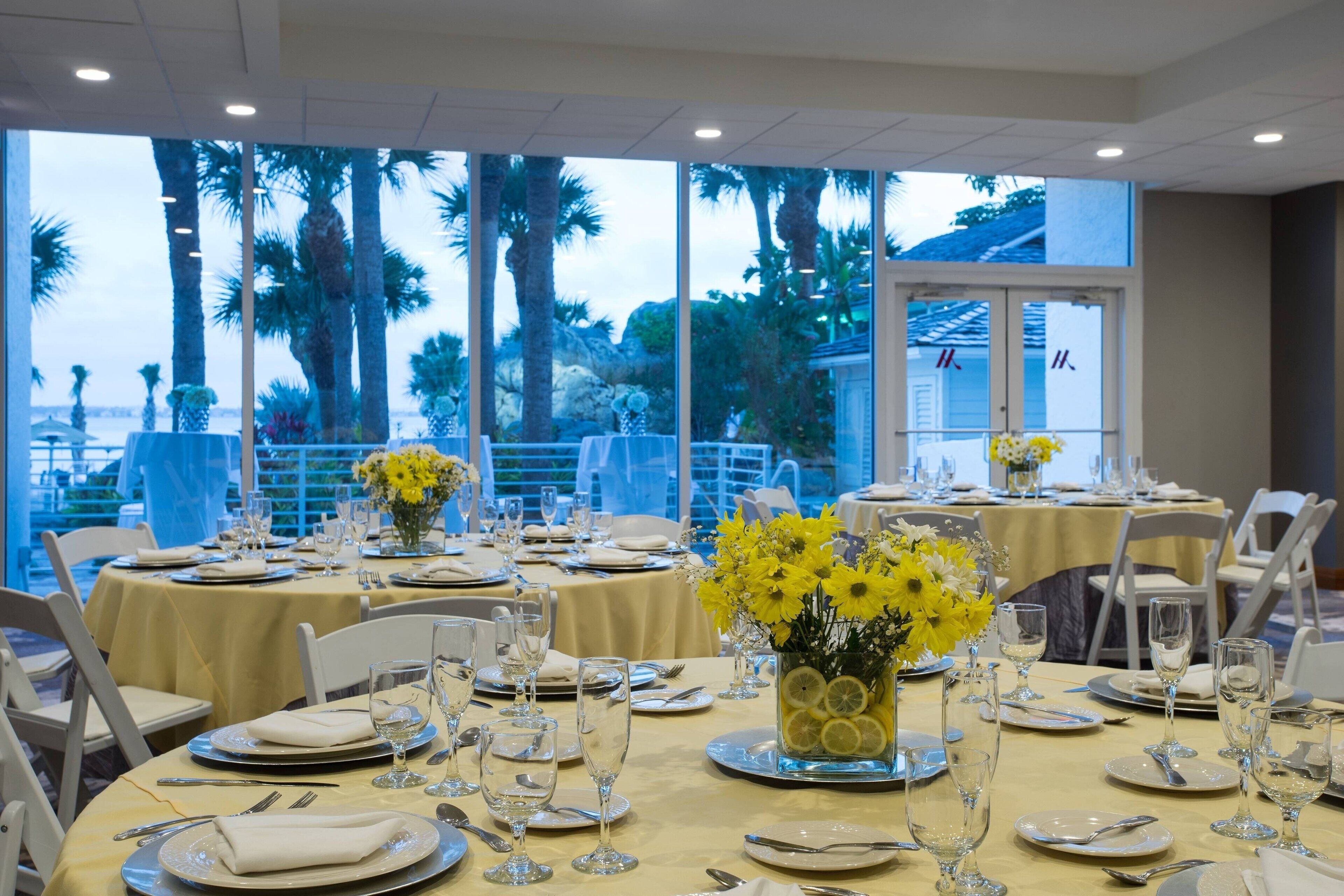 A dining area with tables set for a formal event, featuring yellow flowers and clear glassware.