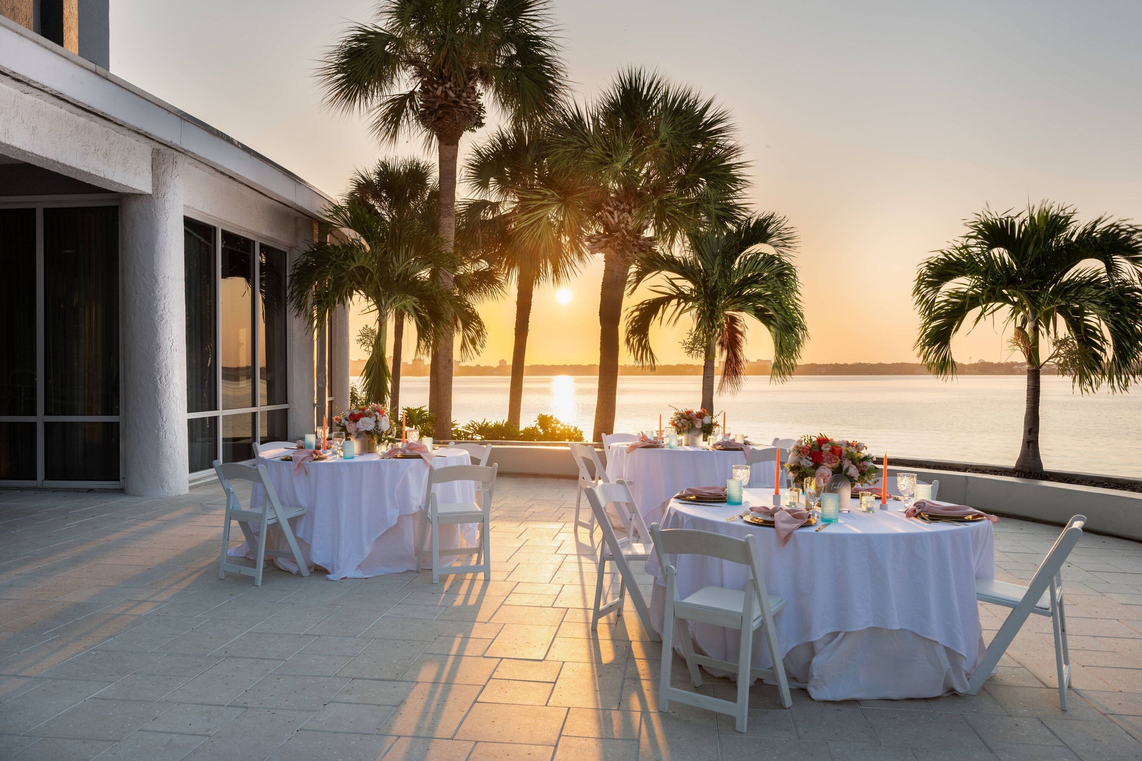 An outdoor dining area with white tables and chairs, set against a sunset view of the ocean.