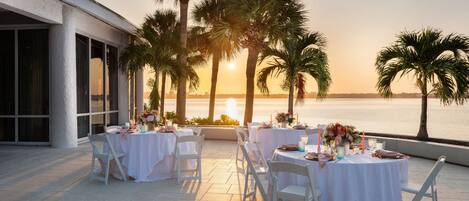 An outdoor dining area with white tables and chairs, set against a sunset view of the ocean.