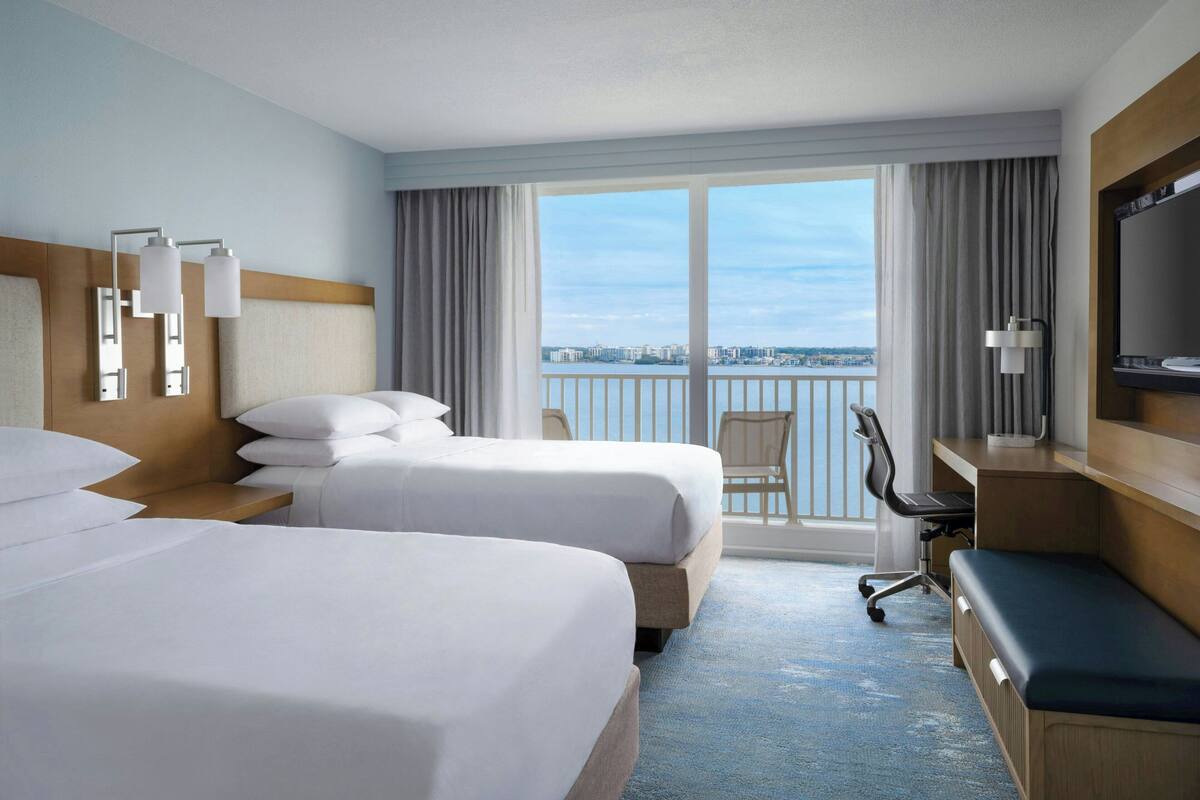 A hotel room with two beds, a desk, a chair, a TV, and a balcony with a city view.