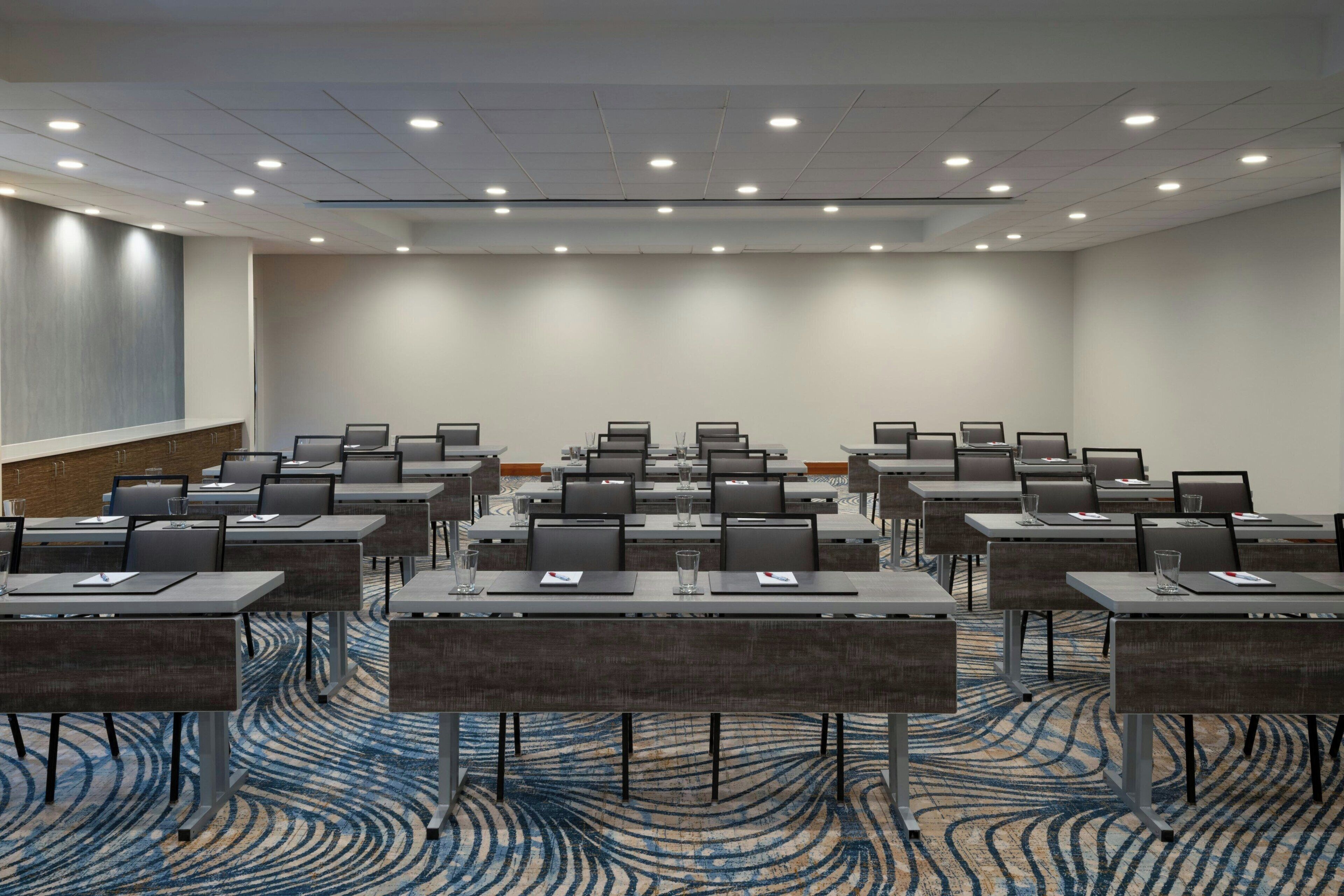 Un salón de conferencias con filas de mesas y sillas, equipado con computadoras portátiles y botellas de agua de vidrio.