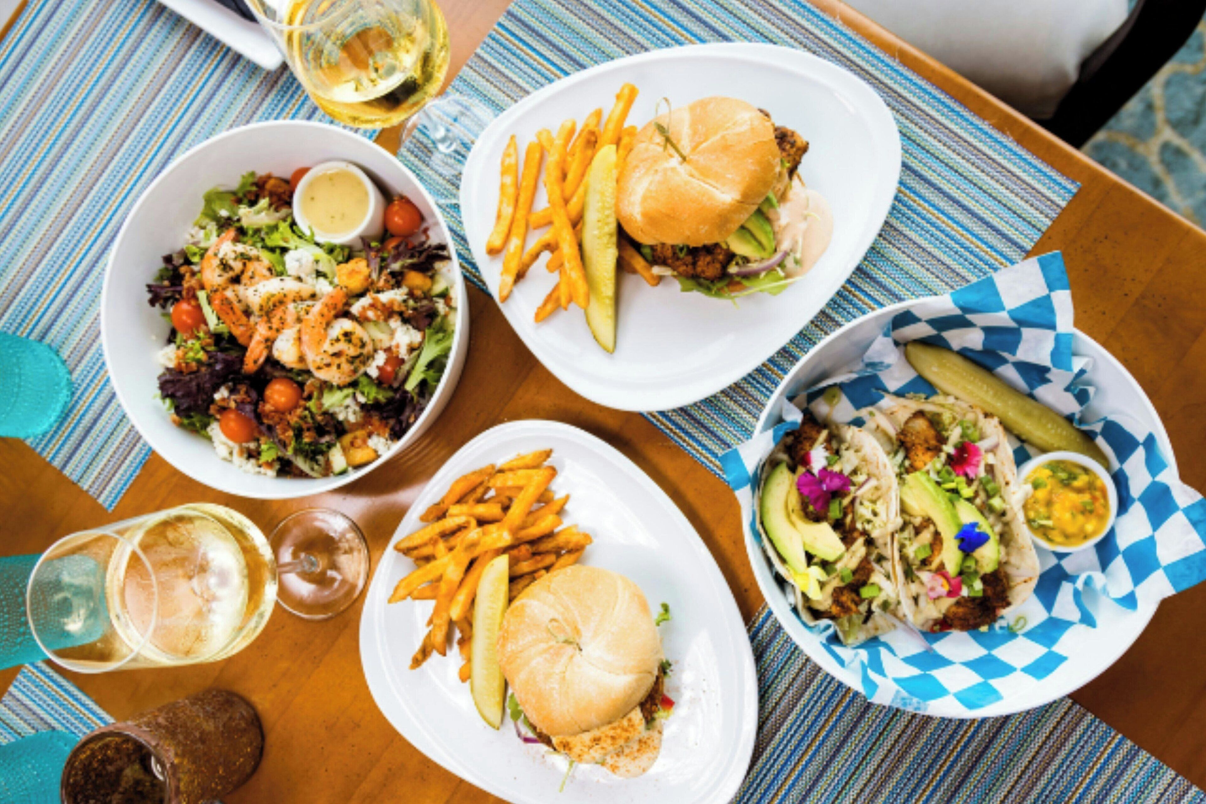 Une table où l’on trouve divers plats, notamment une salade, des hamburgers, des frites et des tacos, ainsi que des verres de vin.