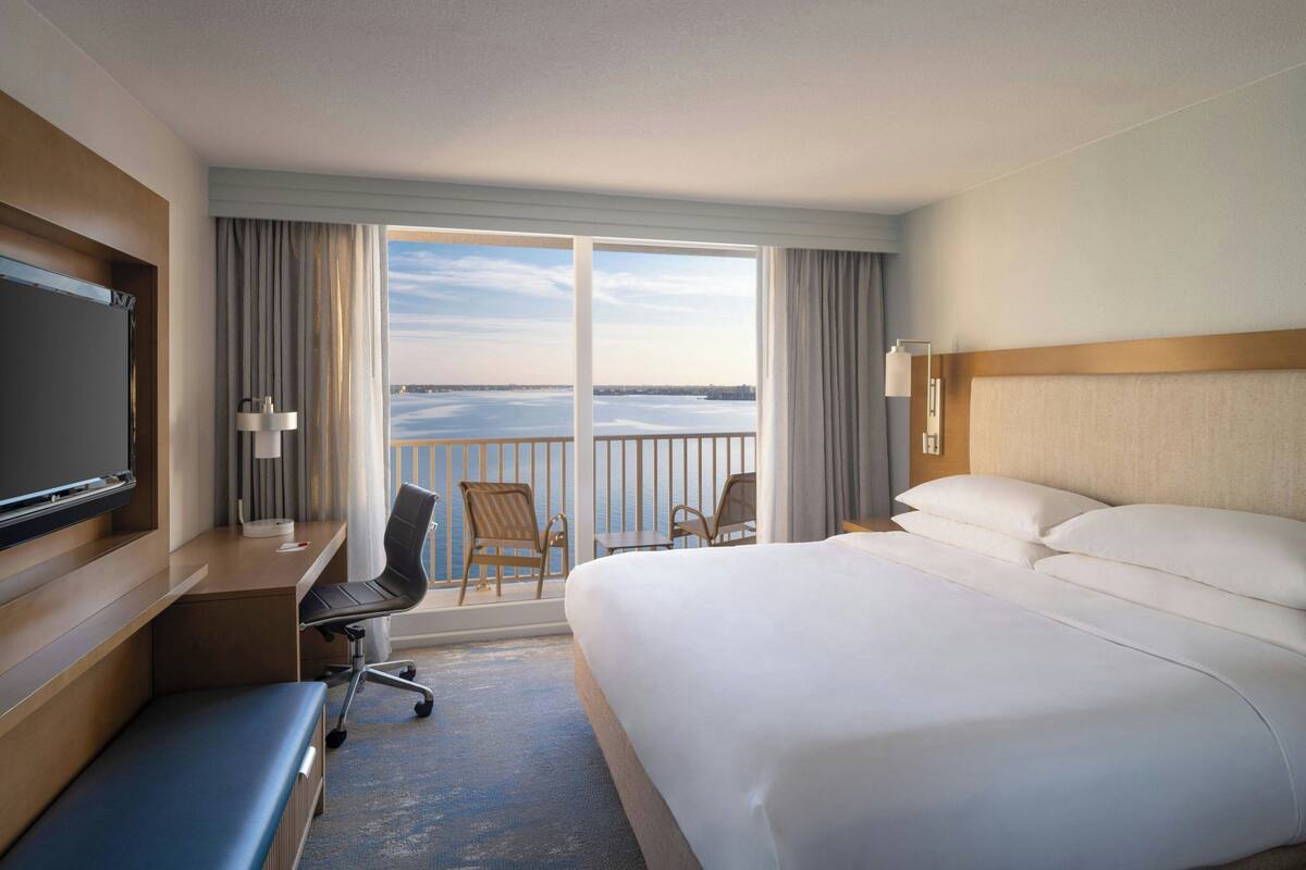 A hotel room with a large bed, a desk with a chair, a television, and a balcony with a view of the water.