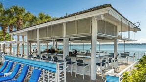 A covered outdoor seating area with lounge chairs and a bar, overlooking a body of water.