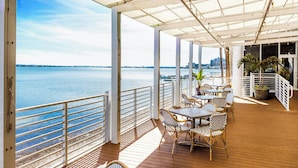 A covered outdoor area with tables and chairs, overlooking a body of water with a distant city skyline.