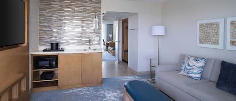 Habitación de hotel moderna con kitchenette, sofá con cojines, banco, televisor de pantalla plana y obras de arte en las paredes.